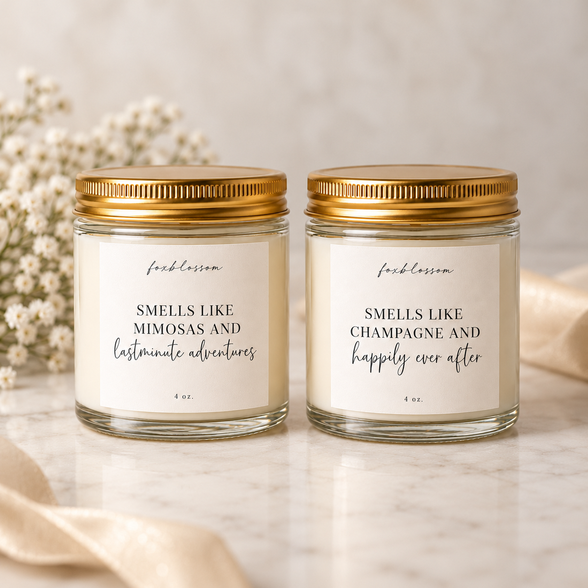 Two 4 oz glass jar candles with gold lids and cream labels, displayed on a marble surface with soft decorative flowers and ribbon. The labels read 'fresh lemon' and 'SMELLS LIKE MIMOSAS AND lastminute adventures' on the left, and 'SMELLS LIKE CHAMPAGNE and happily ever after' on the right.