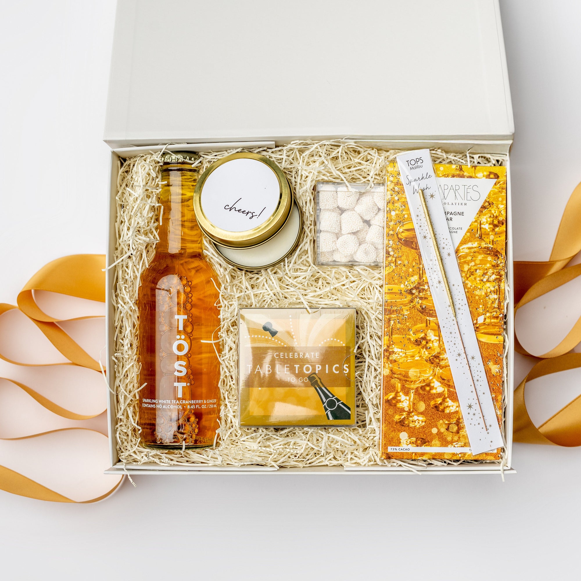 A top view of the Celebration Gift Box featuring a bottle of TÖST Sparkling White Tea, Champagne Bubble Gummies, a Champagne Dark Chocolate Bar, a gold wish sparkler wand, a 4 oz. hand-poured “Cheers” candle, and a deck of Celebrate Conversation Starter Cards, all arranged in a white gift box with ivory shred on a light background and orange ribbon underneath and surrounding the box. 