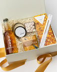 A top view of the Celebration Gift Box featuring a bottle of TÖST Sparkling White Tea, Champagne Bubble Gummies, a Champagne Dark Chocolate Bar, a gold wish sparkler wand, a 4 oz. hand-poured “Cheers” candle, and a deck of Celebrate Conversation Starter Cards, all arranged in a white gift box with ivory shred and tied with a orange ribbon on a light background.