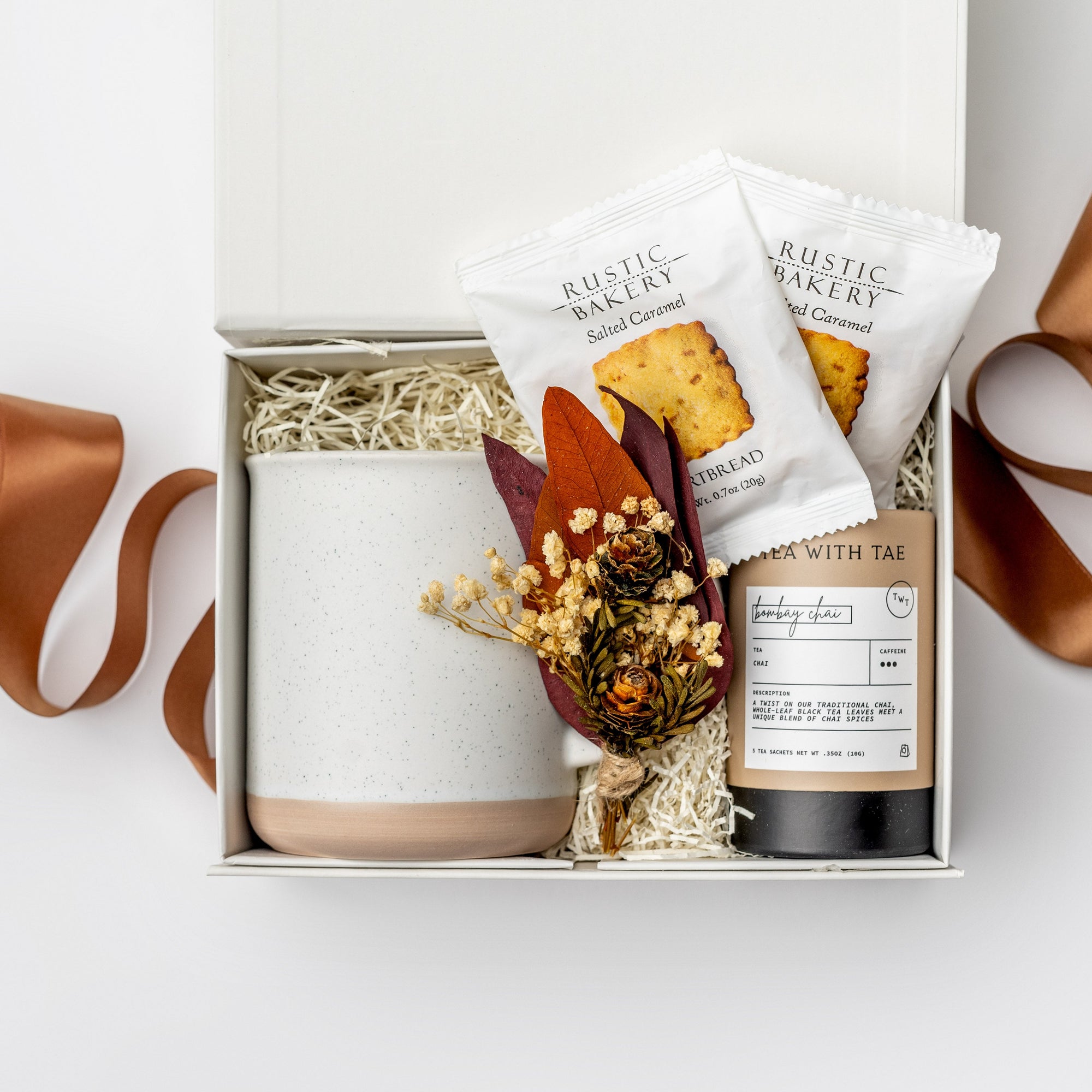 A top view of the Chai Tea Time Gift Box featuring a 12 oz. speckled ceramic mug on the left, an artisan chai tea mini tube and two toffee shortbread cookies in the center, and a handmade dried floral mini bouquet on the right. Arranged in a white gift box with ivory shred and accented with a toffee ribbon crossing underneath the box on a white background.
