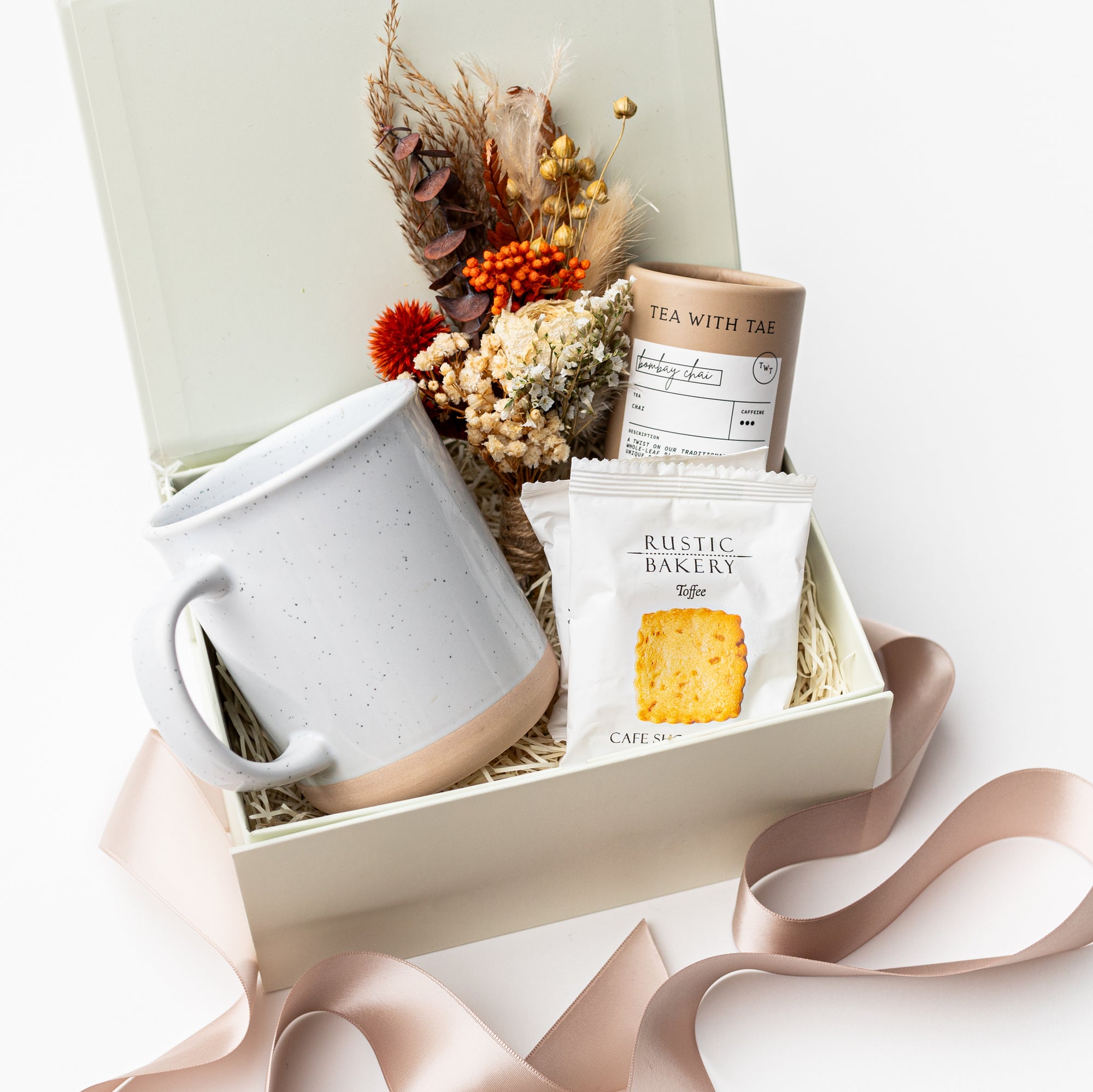 A front view of the Chai Tea Time Gift Box featuring, from left to right, a 12 oz. speckled ceramic mug, an artisan chai tea mini tube, two toffee shortbread cookies, and a handmade dried floral mini bouquet. Presented in a white gift box filled with ivory shred and wrapped with a beige ribbon around the box on a white background.