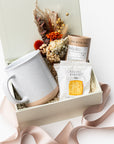 A front view of the Chai Tea Time Gift Box featuring, from left to right, a 12 oz. speckled ceramic mug, an artisan chai tea mini tube, two toffee shortbread cookies, and a handmade dried floral mini bouquet. Presented in a white gift box filled with ivory shred and wrapped with a beige ribbon around the box on a white background.