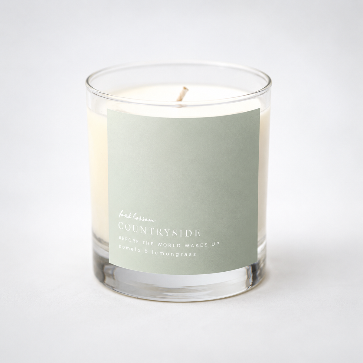 A glass jar candle with cream-colored wax and a single wick, featuring a muted sage green label that reads 'fresh laurel COUNRSIDE' and 'BEFORE THE WORLD WAKES UP'.