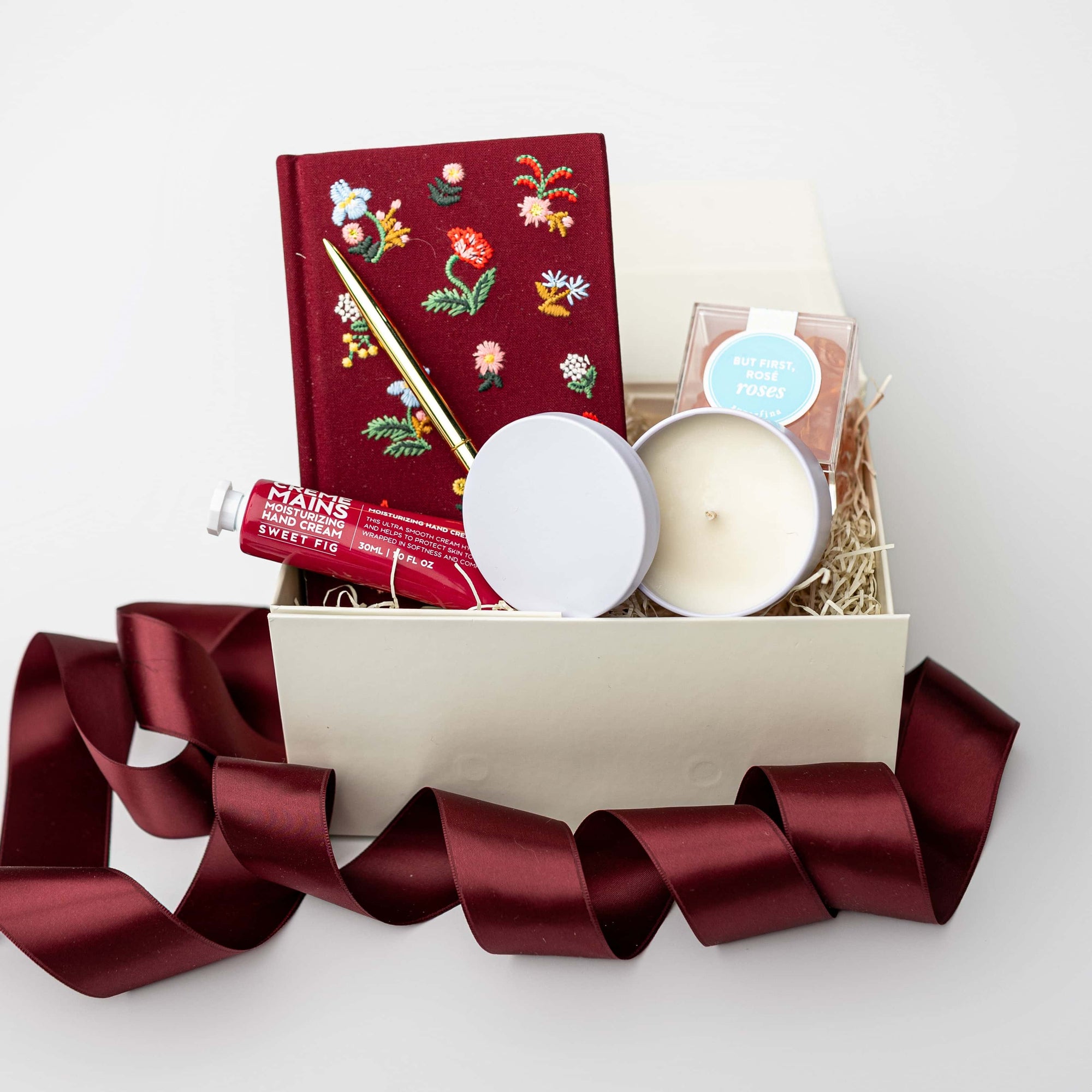 Open gift box filled with shredded paper containing a burgundy floral embroidered journal with a gold pen, hand cream, and a small candle jar, styled on a white background with burgundy ribbon accents.