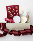 Open gift box filled with shredded paper containing a burgundy floral embroidered journal with a gold pen, hand cream, and a small candle jar, styled on a white background with burgundy ribbon accents.
