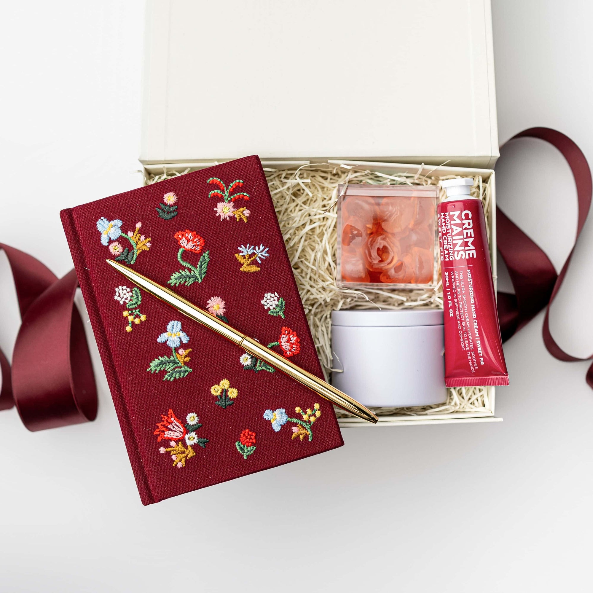 Open gift box filled with shredded paper containing a burgundy floral embroidered journal with a gold pen, hand cream, and a small candle jar, styled on a white background with burgundy ribbon accents.