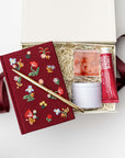 Open gift box filled with shredded paper containing a burgundy floral embroidered journal with a gold pen, hand cream, and a small candle jar, styled on a white background with burgundy ribbon accents.