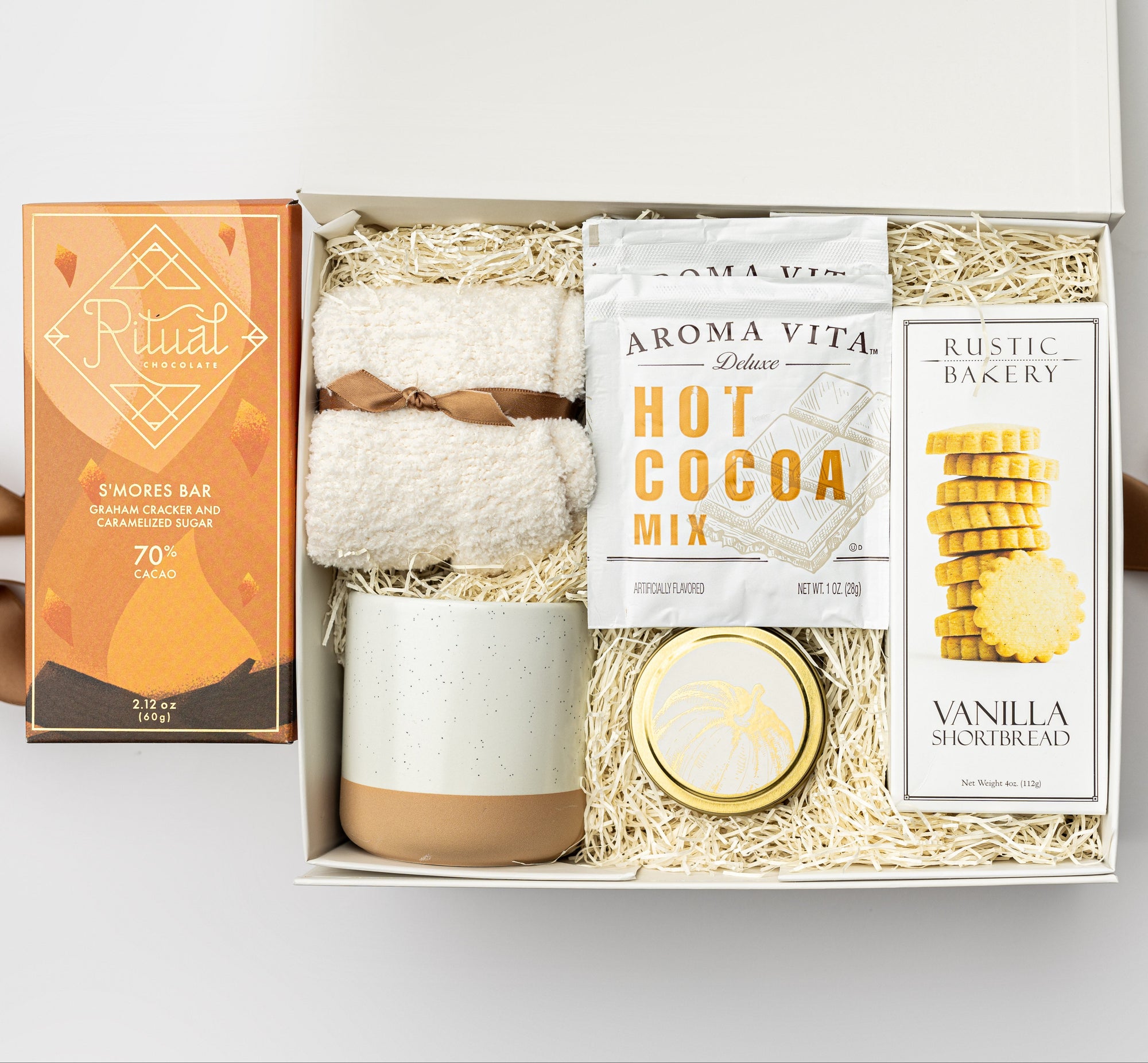A top view of a cozy fall gift set featuring cloud socks, vanilla shortbread cookies, a S’mores cacao bar, a 4 oz. pumpkin spice candle, a set of two single-serving hot cocoa packets, and a speckled ceramic mug, all arranged in a white gift box with ivory shred and tied with a warm orange ribbon on a white background.