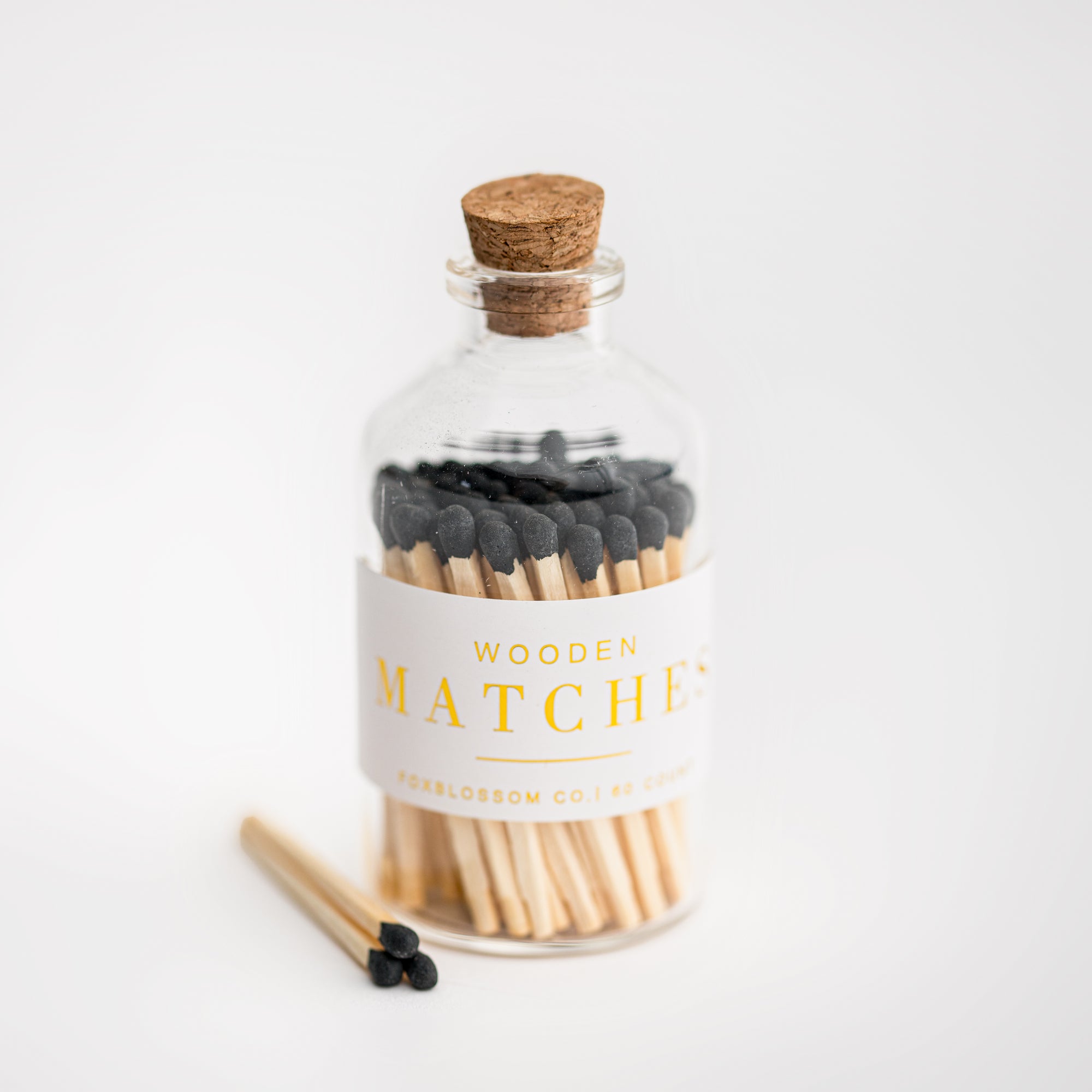 Foxblossom custom black tip matches are featured in a corked glass jar with a white label with gold lettering on a white background with three matches outside of the jar. 