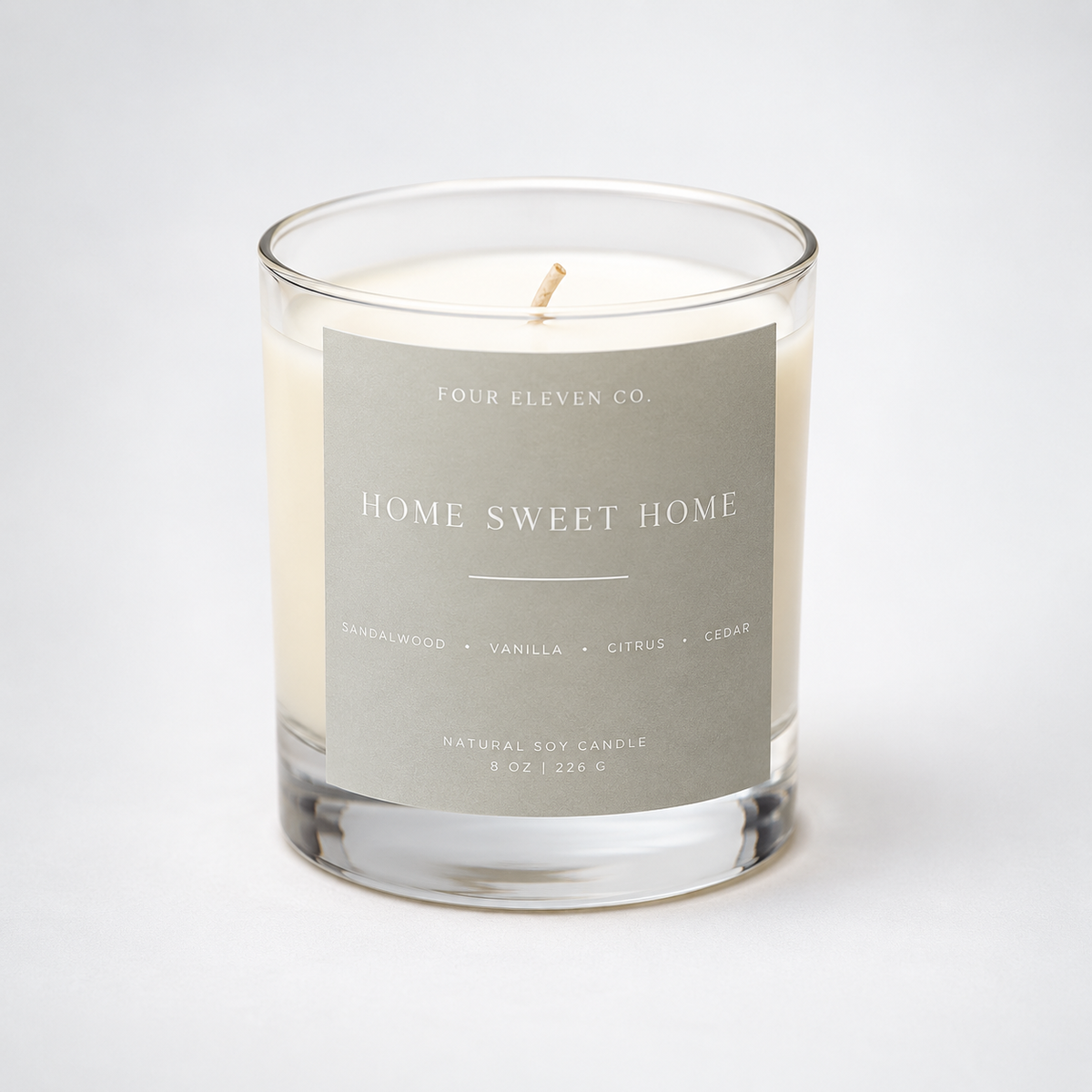 A clear glass jar candle with white wax and a single wick, featuring a light gray label that reads 'FOUR ELEVEN CO. HOME SWEET HOME' on a clean white background.