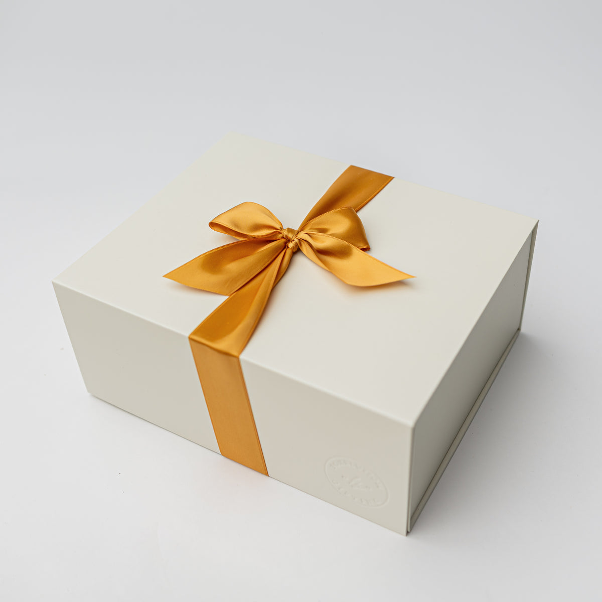 A cream-colored rectangular gift box tied with a satin gold ribbon and bow, photographed on a clean light gray background.