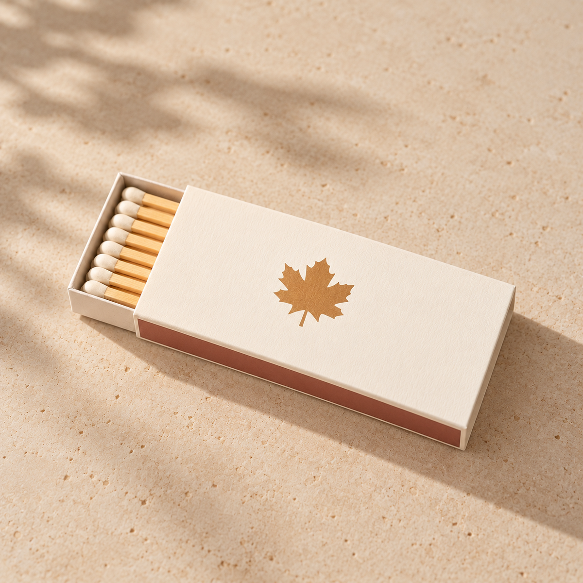 An open matchbox with wooden matches inside, placed on a light beige textured surface. The matchbox lid is off-white with a gold maple leaf emblem, and the side of the box is a muted dusty rose.