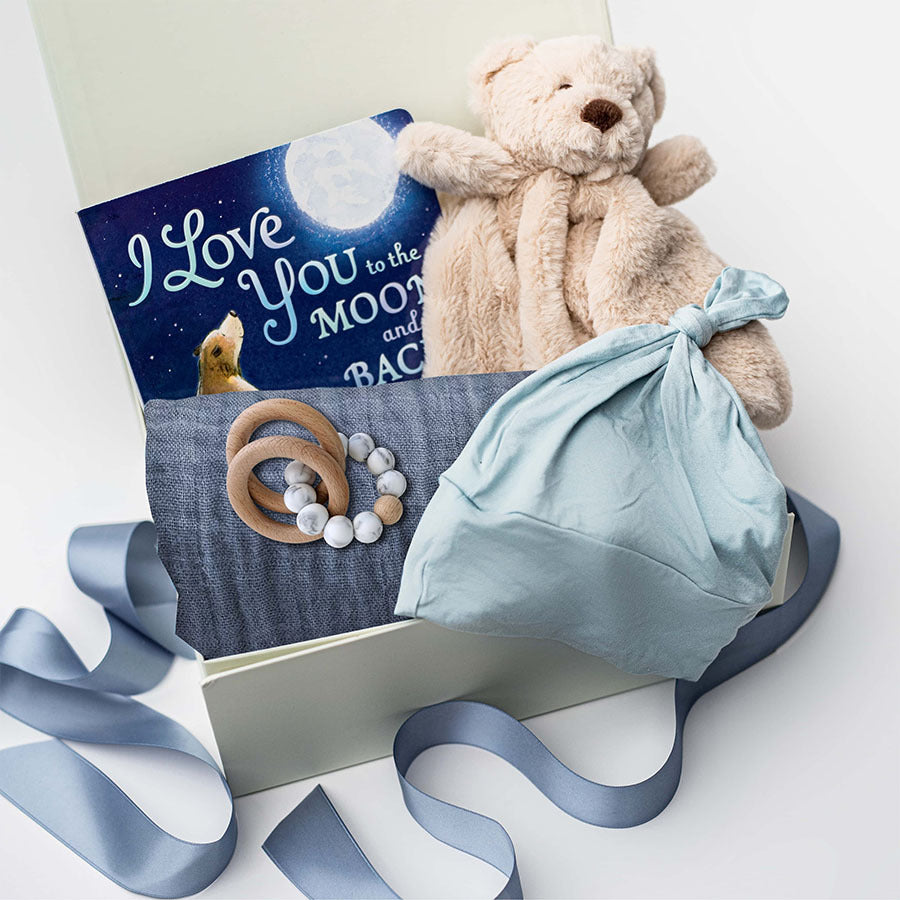 A white gift box on a white background, filled with light ivory paper shred and tied with a soft blue ribbon. Inside the box: a plush teddy bear lovey (with optional personalization), a muslin cotton swaddle blanket printed with white stars on a dusty-blue background, a soft blue stretch baby top-knot hat, a silicone and wood baby teether consisting of two wooden rings and a grey-and-white bead loop, and a board book titled “I Love You to the Moon and Back.”