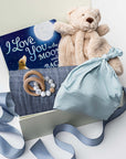 A white gift box on a white background, filled with light ivory paper shred and tied with a soft blue ribbon. Inside the box: a plush teddy bear lovey (with optional personalization), a muslin cotton swaddle blanket printed with white stars on a dusty-blue background, a soft blue stretch baby top-knot hat, a silicone and wood baby teether consisting of two wooden rings and a grey-and-white bead loop, and a board book titled “I Love You to the Moon and Back.”