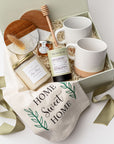 A top view of the New Home Housewarming Gift Box featuring a set of two marble and wood coasters, a “Home Sweet Home” tea towel, a 9 oz. hand-poured candle labeled “New Beginnings, New Home, New Memories” (optionally personalized with a family name), a mini tea tube of jasmine green tea, a mini jar of honey with a wooden honey drizzler, and a set of two speckled stoneware mugs, all arranged in a white gift box with ivory shred and tied with a green ribbon on a white background.