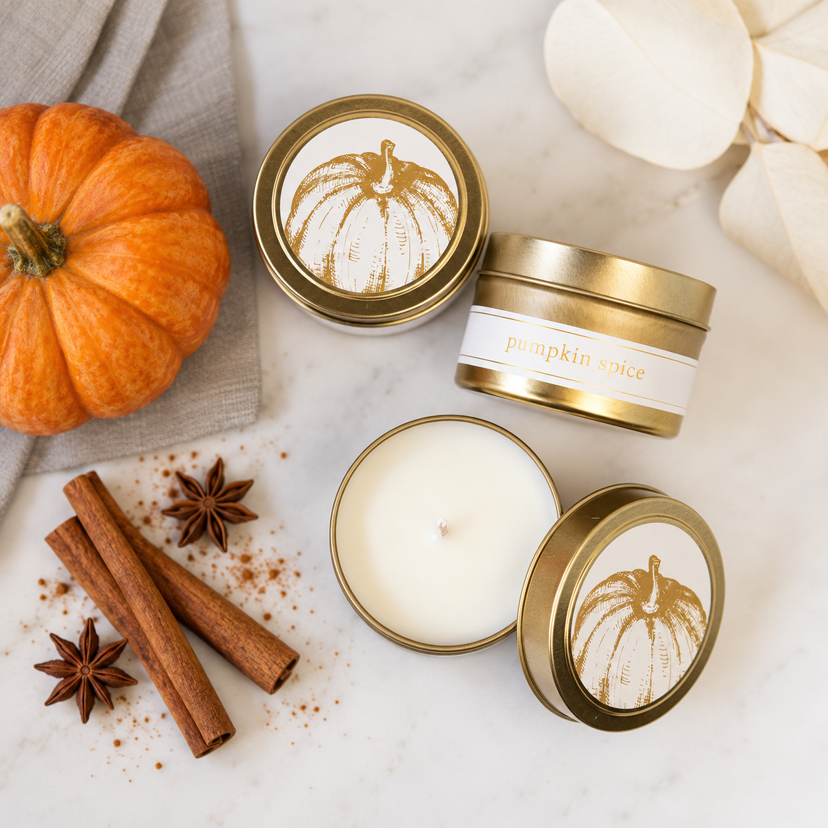 Three gold tin candles with white labels featuring a pumpkin illustration are arranged on a white marble surface with autumn props including a small orange pumpkin, cinnamon sticks, star anise, and cream petals. One candle is open, showing white wax, while the others are closed.