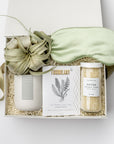 A top view of a soothing spa-themed gift box presented in a white gift box filled with ivory shred and tied with a green ribbon, featuring detox bath salts, eucalyptus & peppermint sea mineral foot soak, a 12 oz. hand-poured eucalyptus & lavender candle in a matte white ceramic jar, a Tillandsia Xerographica live air plant, and a sage silk sleep mask, all arranged on a white background.