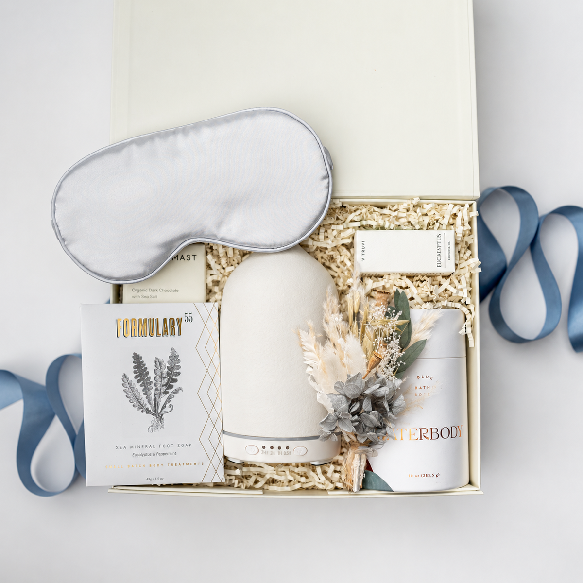 A curated gift box set on a white background, featuring a white sleep mask, a white diffuser, a white candle, a card from Formulary, and a small soap, all arranged in a cream gift box with shredded paper and blue ribbon accents.