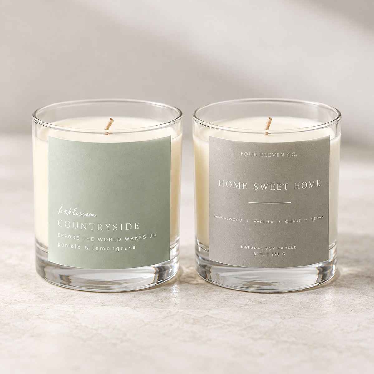 Two lit scented candles in clear glass jars on a light stone surface. The left candle has a pale sage green label reading 'bubbleum Countryside,' and the right candle has a light gray label reading 'FOUR ELEVEN CO.