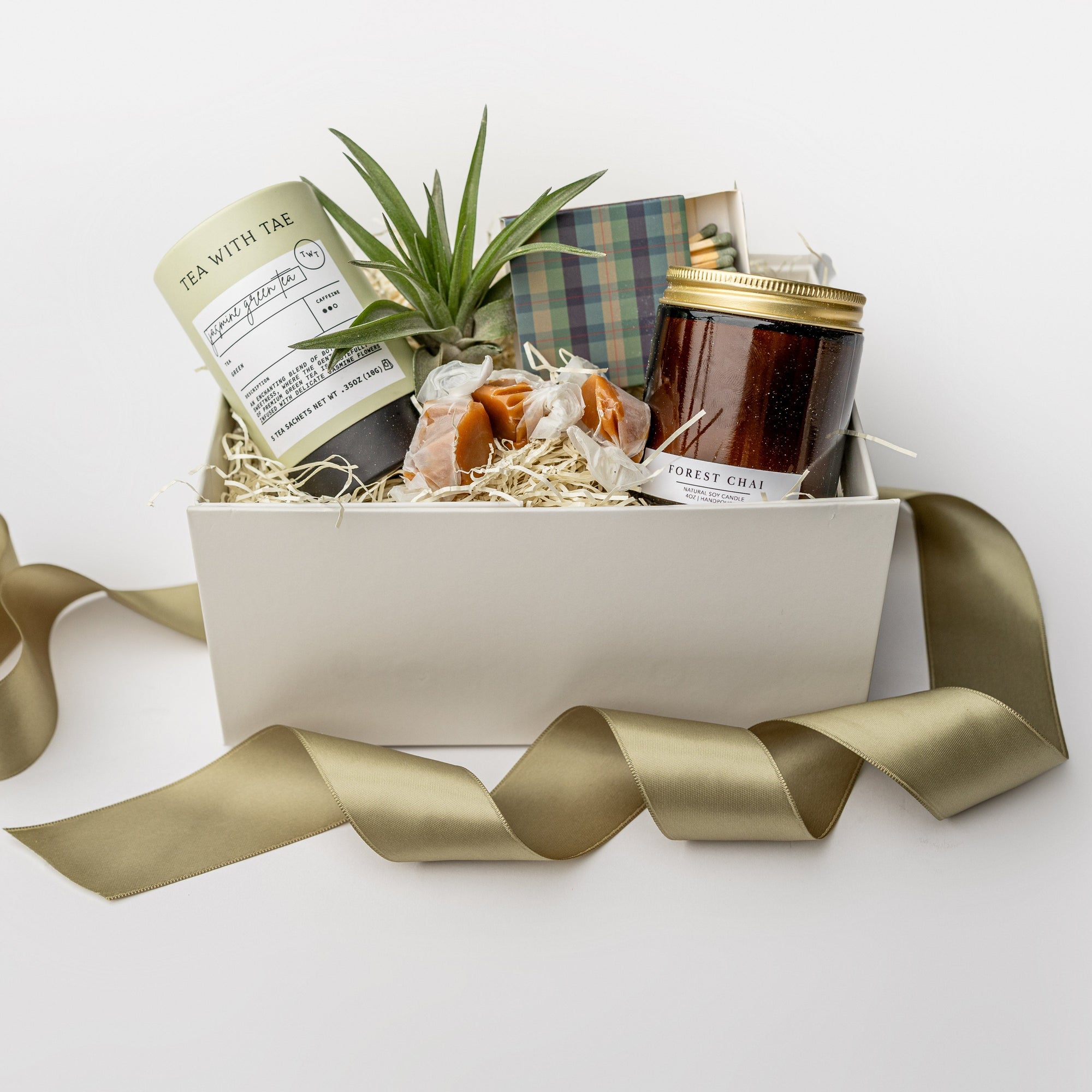 A white gift box with ivory paper shred and a green ribbon on a white background. Inside the box are a small live air plant with a care card, a mini tube of artisan jasmine green tea, a 4oz hand-poured soy candle in the scent Forest Chai, a matchbox of navy and green plaid-patterned matches, and three loose caramels — a thoughtful “Sending Thanks” gift box.