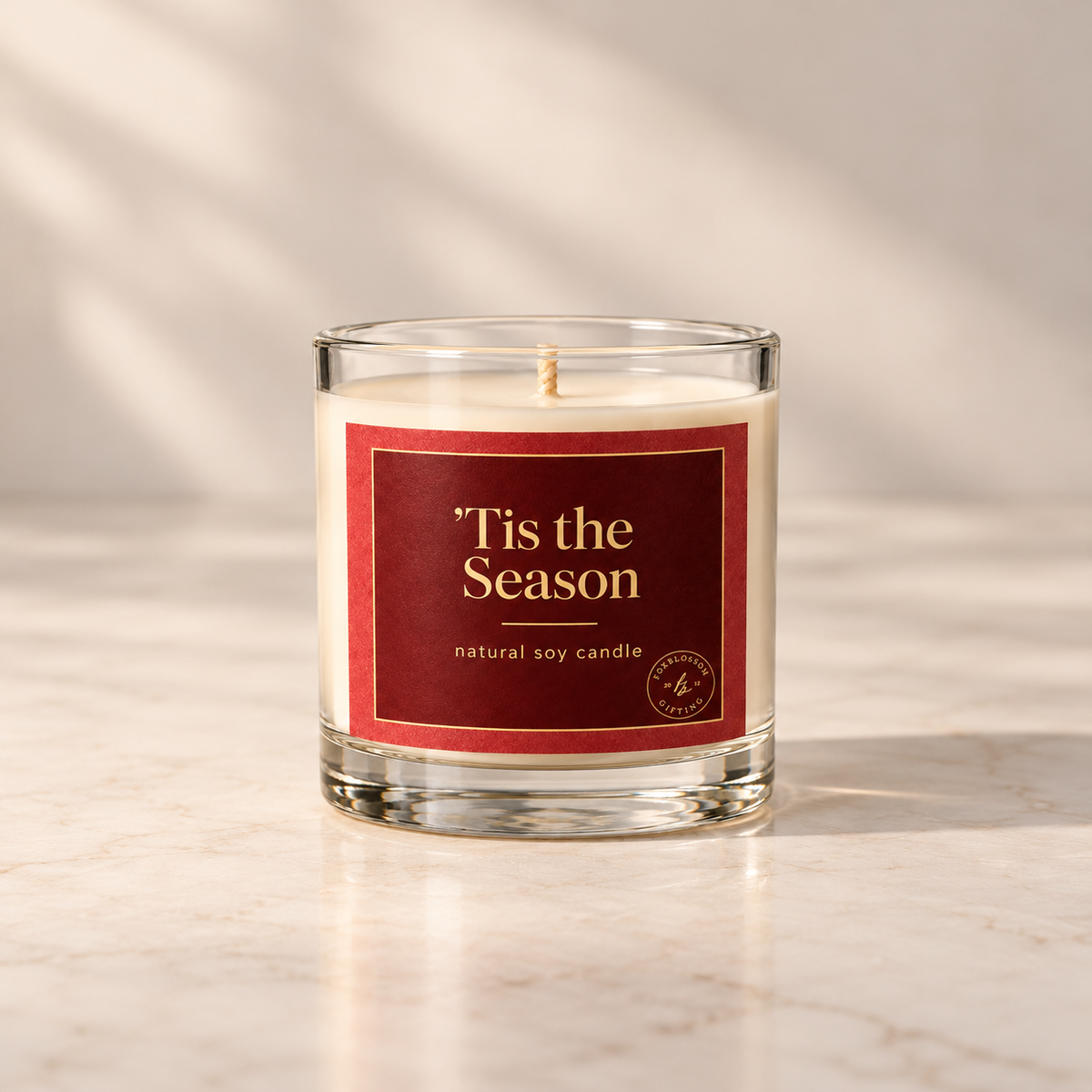 A clear glass jar candle with cream-colored wax and a red label reading '*Tis the Season' and 'natural soy candle,' set on a light marble surface with soft shadow overlays.