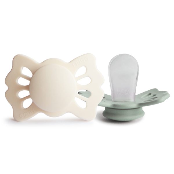 The image shows two baby pacifiers with butterfly-shaped shields, one cream-colored and the other a muted green, set against a clean white background.