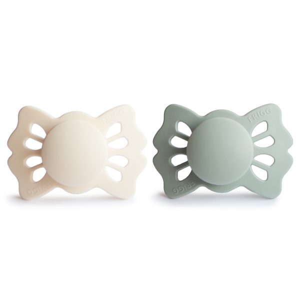 The image shows two baby pacifiers with butterfly-shaped shields, one cream-colored and the other a muted green, set against a clean white background.
