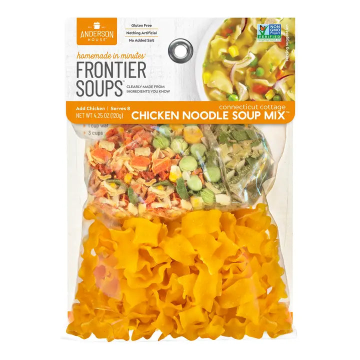 Frontier chicken noodle soup mix is featured a clear plastic bag, divided into sections containing dried soup ingredients. The bottom section is filled with yellow, ruffled egg noodles. Above the noodles, the bag is sectioned into three, showing a mix of dried vegetables, including orange carrots, green peas, and other herbs and spices. A small picture of the prepared soup in the label with black and white lettering has a silver ring attached on a white background.  