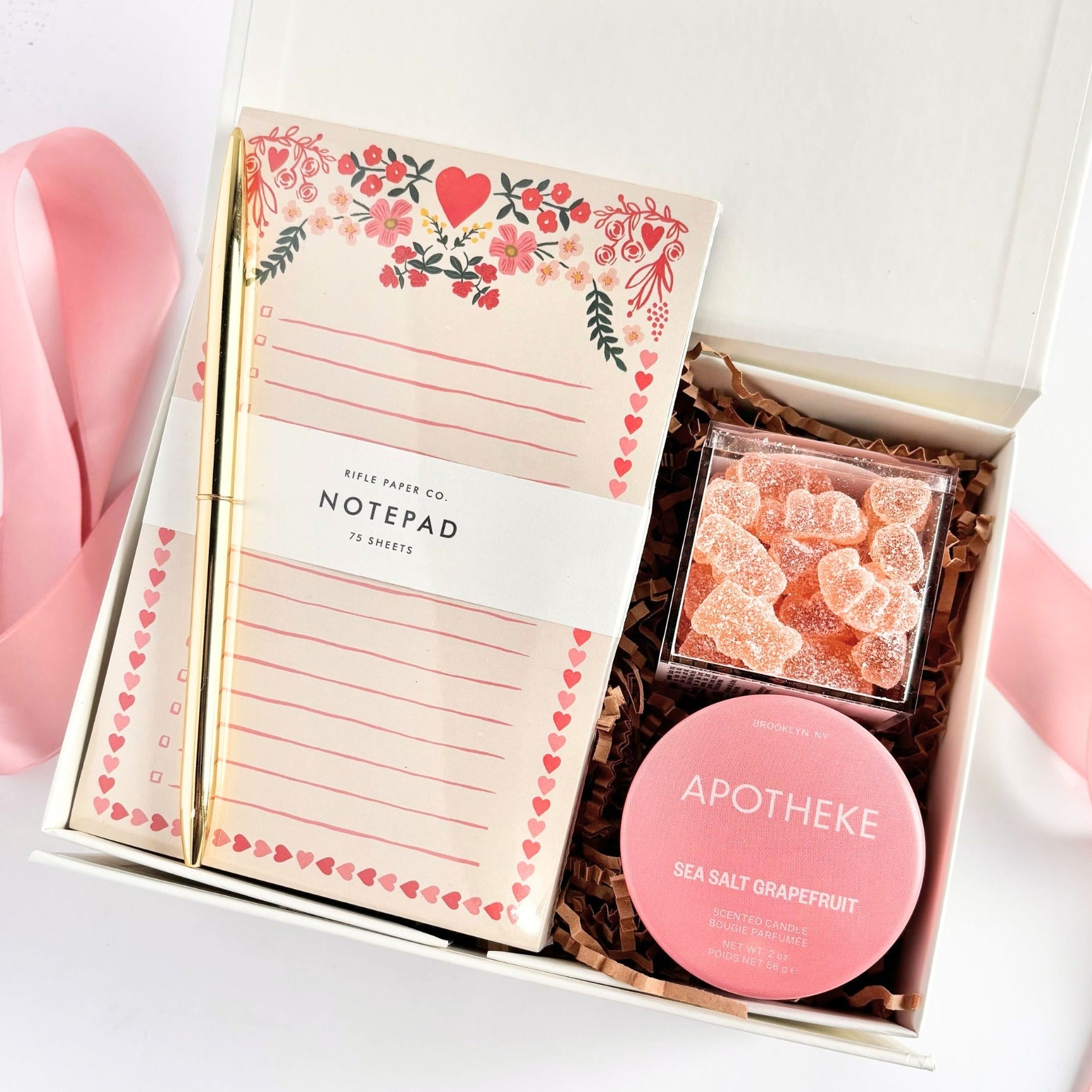 White gift box with brown crinkle paper filled with a floral-heart printed notepad, a gold pen, a clear square container of pink sugar-coated gummy candies, and a round pink Apotheke sea salt grapefruit scented candle, with a pink ribbon beside the box.