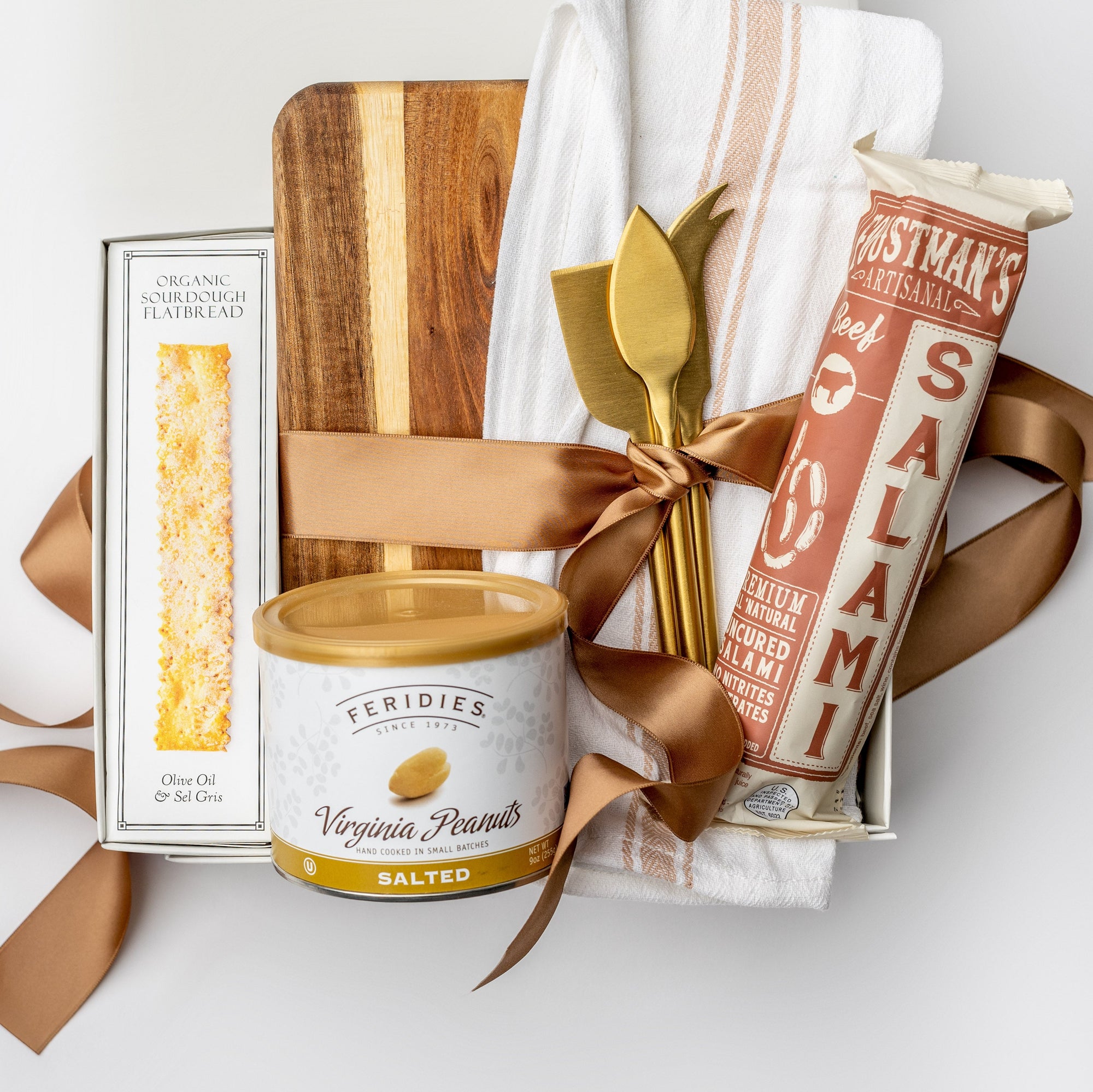 A white gift box with ivory paper shred and a brown ribbon on a white background. Inside the box are an artisan pork all-natural uncured smoked salami, everything spice flatbread crackers, a 9oz jar of sea salt and black pepper peanuts, a neutral striped tea towel, a set of three gold-plated cheese knives, a walnut charcuterie board (optionally engraved), and a small dried bunny tail decoration — an elegant gourmet charcuterie gift set.