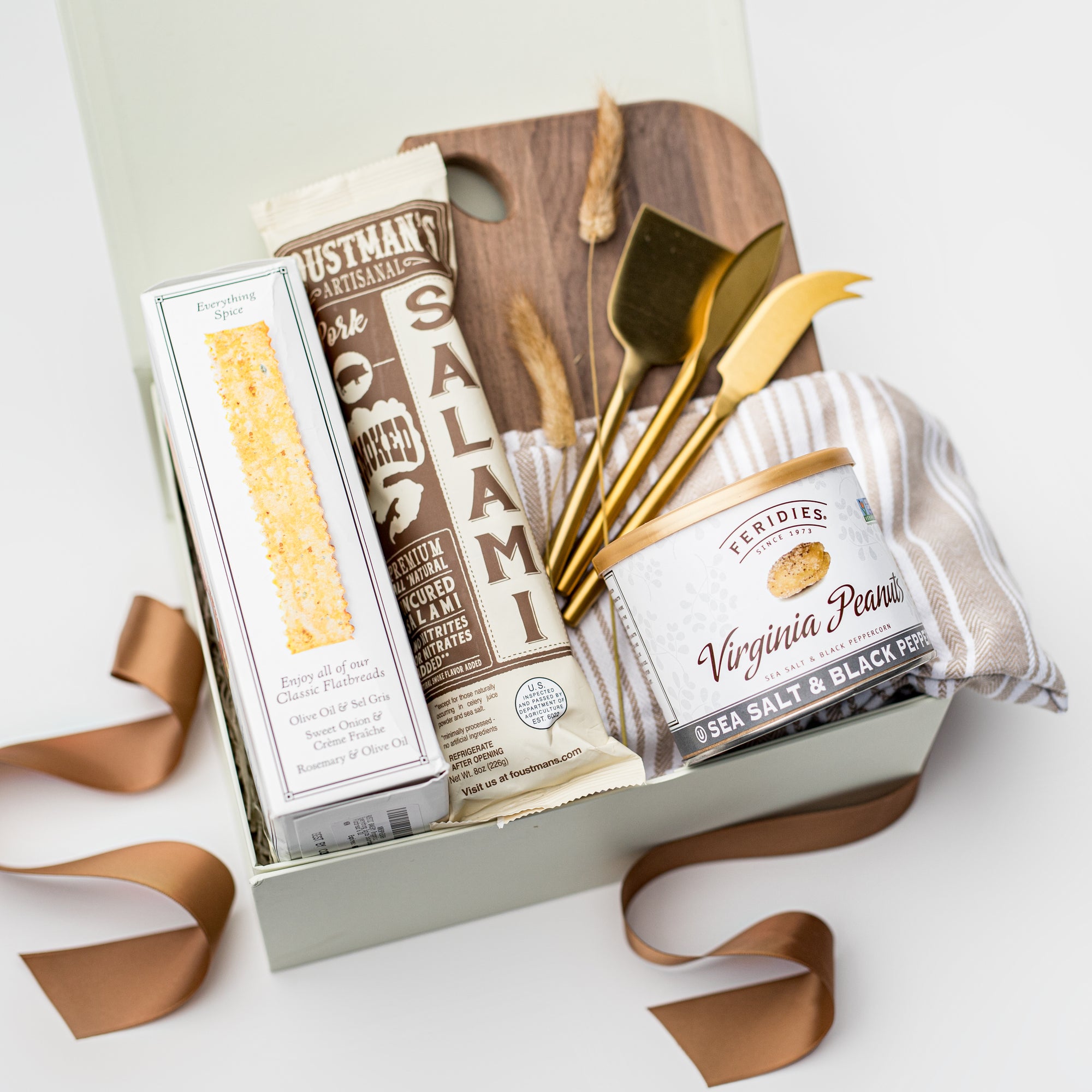 A white gift box with ivory paper shred and a brown ribbon on a white background. Inside the box are an artisan pork all-natural uncured smoked salami, everything spice flatbread crackers, a 9oz jar of sea salt and black pepper peanuts, a neutral striped tea towel, a set of three gold-plated cheese knives, a walnut charcuterie board (optionally engraved), and a small dried bunny tail decoration — an elegant gourmet charcuterie gift set.