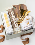 A white gift box with ivory paper shred and a brown ribbon on a white background. Inside the box are an artisan pork all-natural uncured smoked salami, everything spice flatbread crackers, a 9oz jar of sea salt and black pepper peanuts, a neutral striped tea towel, a set of three gold-plated cheese knives, a walnut charcuterie board (optionally engraved), and a small dried bunny tail decoration — an elegant gourmet charcuterie gift set.