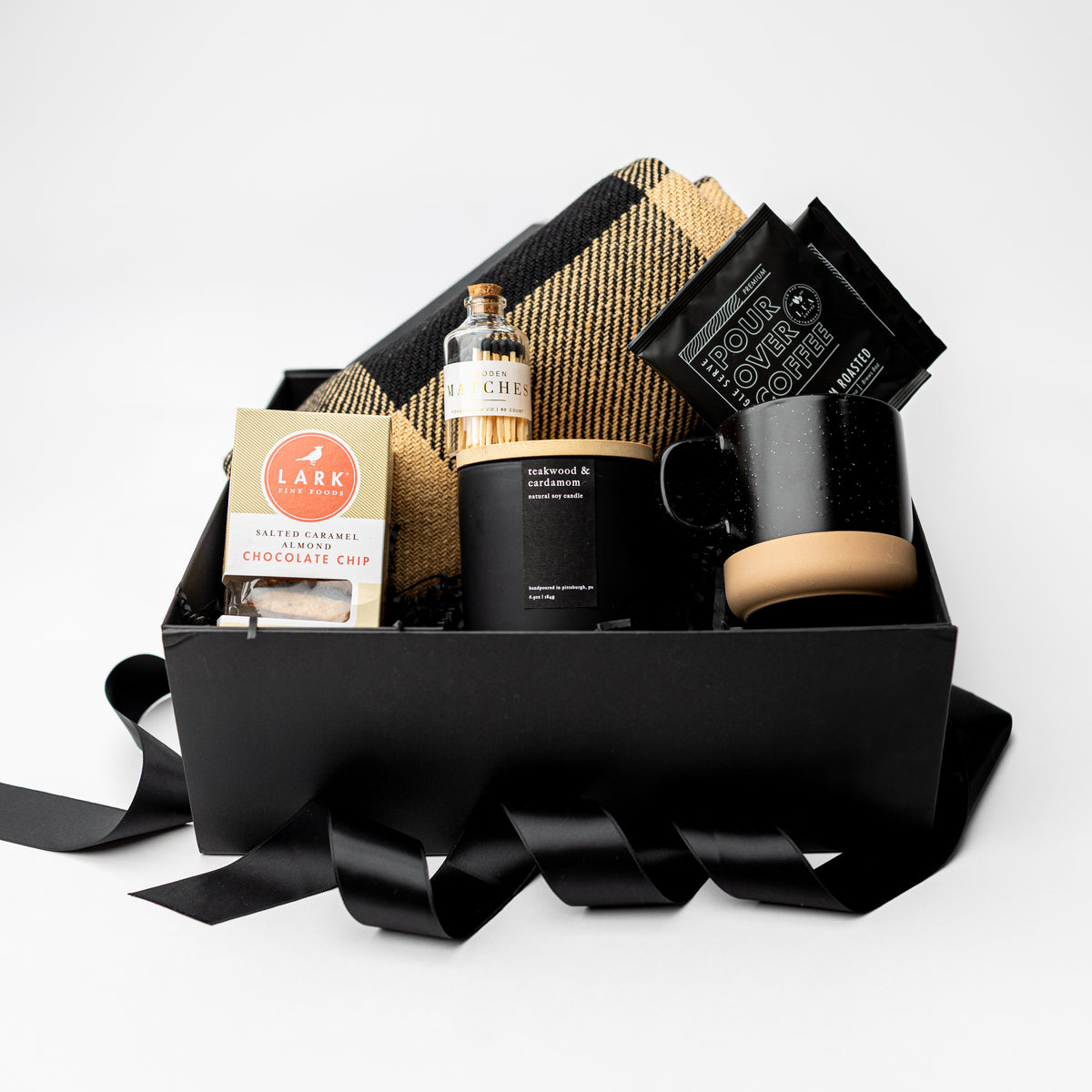 A curated gift box set on a white background, featuring a black gift box with a black ribbon, a black mug with a wooden coaster, a black candle, a bottle of whiskey, chocolate chip cookies, and a black-and-gold plaid blanket.