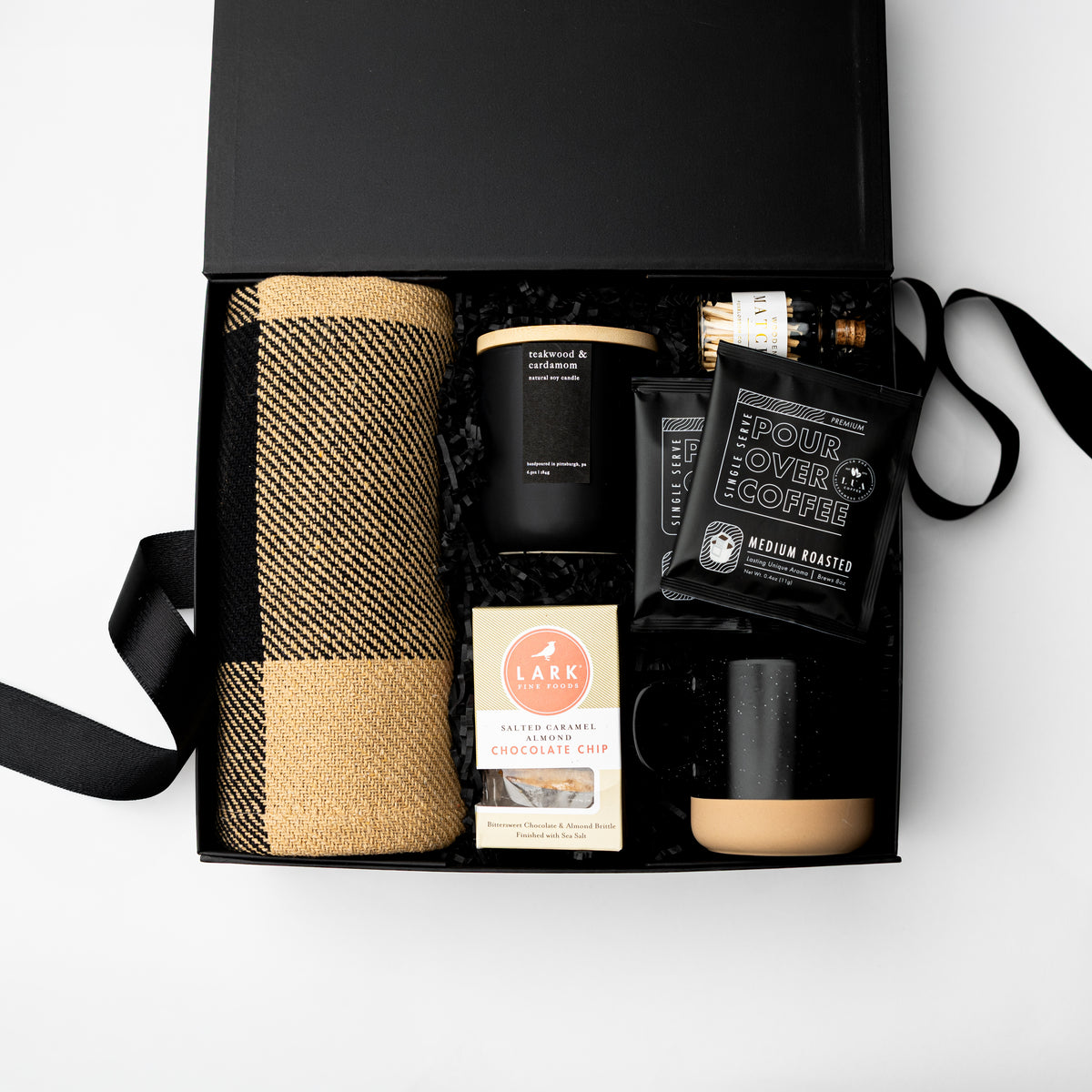 An open black gift box on a white background containing a beige-and-brown plaid blanket, a black candle, a small bottle, a black coffee bag labeled Pour Over Coffee, a Lark box, and a black mug with a beige base.