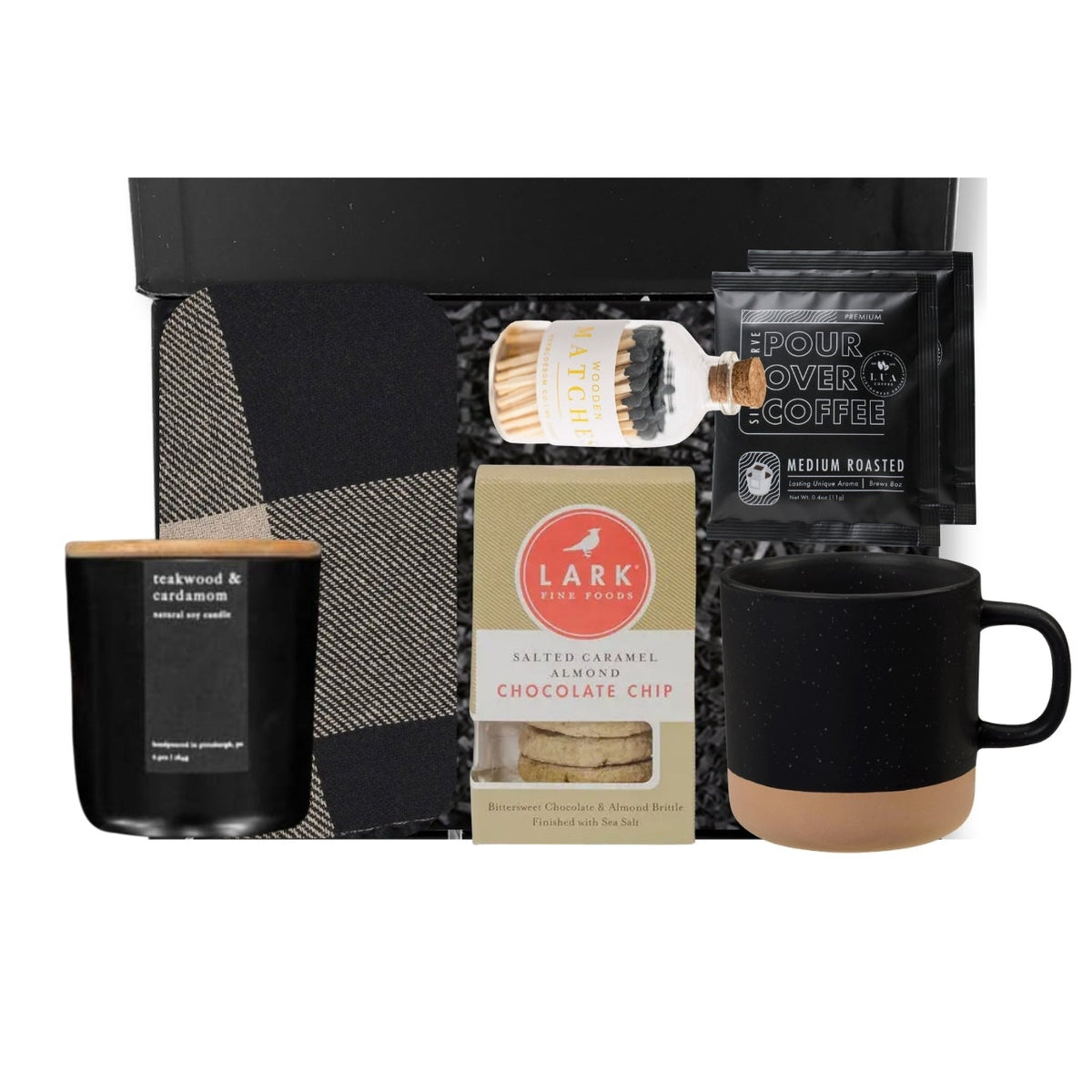 A curated coffee and home gift set arranged on a white background, including a black candle with a light wood lid, a box of Lark Chocolate Chip cookies, a black pour-over coffee bag, a black mug with a tan base, and a black gift box with a plaid pattern.