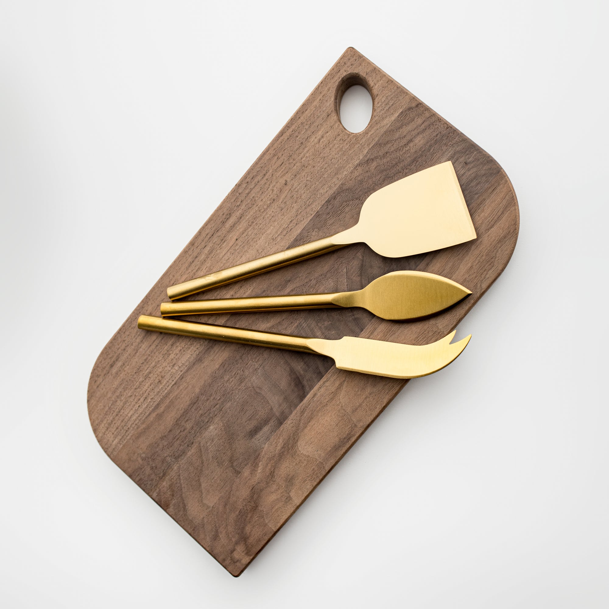 Matte gold cheese knives, set of three, gold-plated stainless steel, one pronged knife for hard cheeses, one wedge knife for semi-hard cheeses, and one tear-shaped knife for soft cheeses. Shown on top of a wooden cheese board in a white background.  