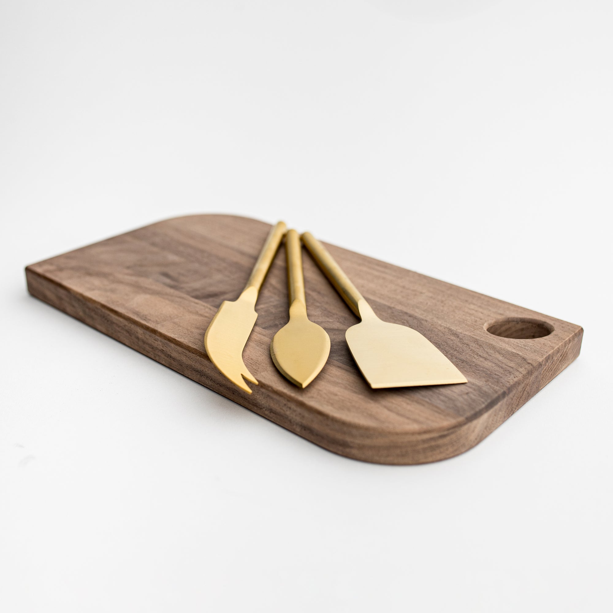 Matte gold cheese knives, set of three, gold-plated stainless steel, one pronged knife for hard cheeses, one wedge knife for semi-hard cheeses, and one tear-shaped knife for soft cheeses. Shown side way on top of a wooden cheese board in a white background.  