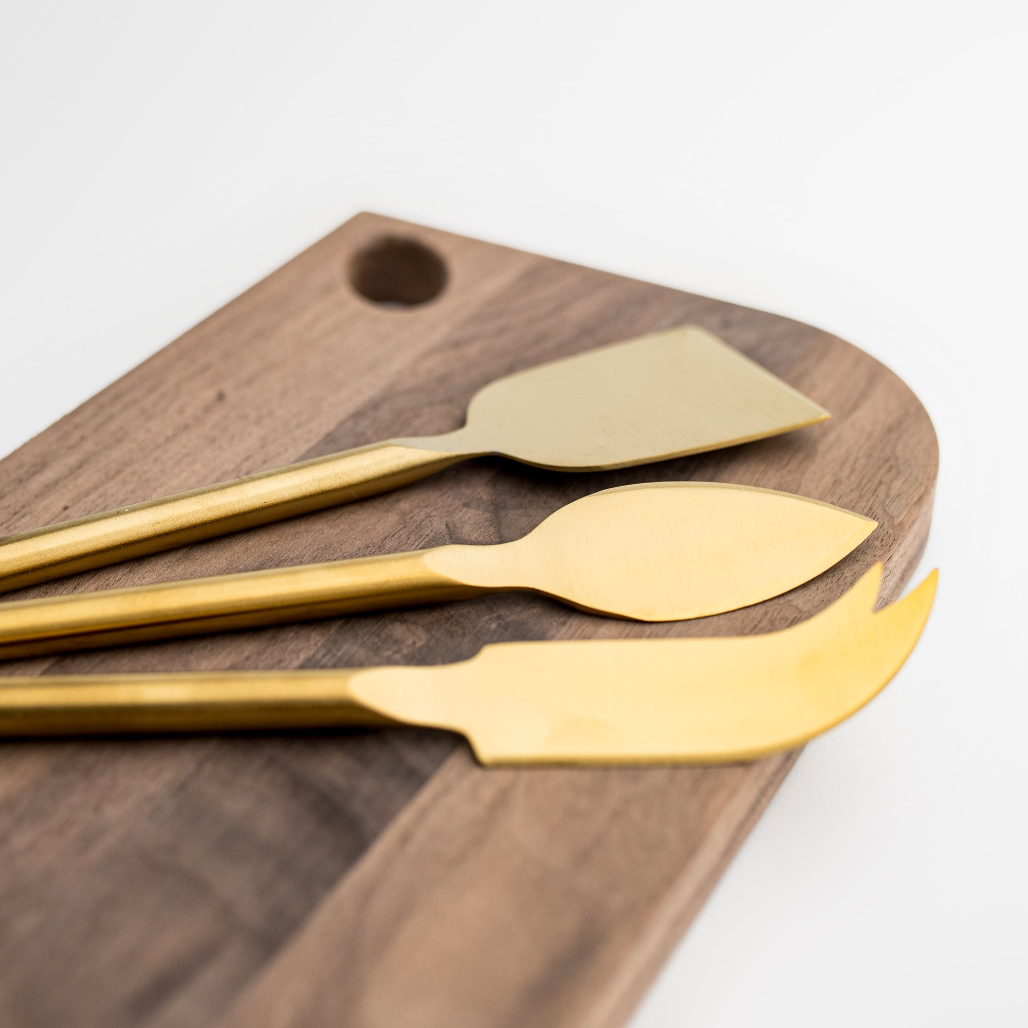 Matte gold cheese knives, set of three, gold-plated stainless steel, one pronged knife for hard cheeses, one wedge knife for semi-hard cheeses, and one tear-shaped knife for soft cheeses. Shown form a corner view on top of a wooden cheese board in a white background.  