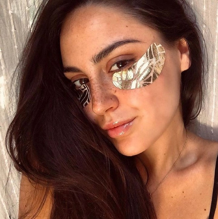 Gold eye pads from Vita Masques is featured a woman with long, dark, slightly wavy hair flowing down her shoulder. She has a fair complexion, with freckles scattered across her nose and cheeks, enhancing her natural beauty. Her eyes are light brown, and her gaze is direct. She wears the golden eye patches with a leaf-like pattern. Her lips are slightly parted, and glossy. The woman's expression is relaxed, with a hint of a smile wearing a simple black top, with a delicate gold chain. In a room background. 