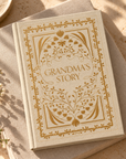A hardcover book titled "Grandma's Story" with ornate gold floral border artwork on an off-white cover, styled on a linen surface with warm sunlight and small white flowers nearby.
