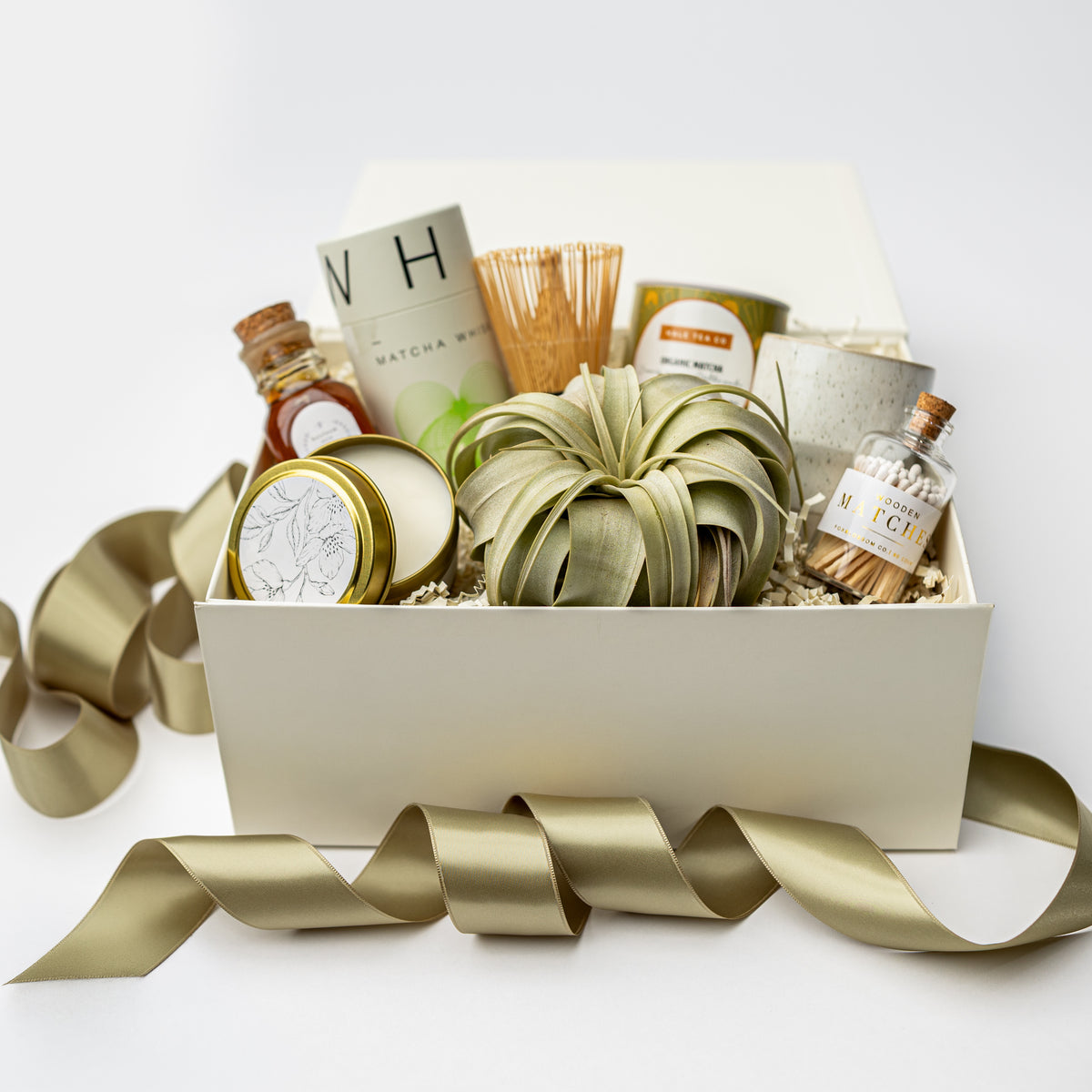 A curated gift box filled with assorted home and wellness items, including a green decorative bundle, small bottles, candles, and a bamboo whisk, arranged in a white box with a gold ribbon on a white background.