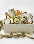 A curated gift box filled with assorted home and wellness items, including a green decorative bundle, small bottles, candles, and a bamboo whisk, arranged in a white box with a gold ribbon on a white background.