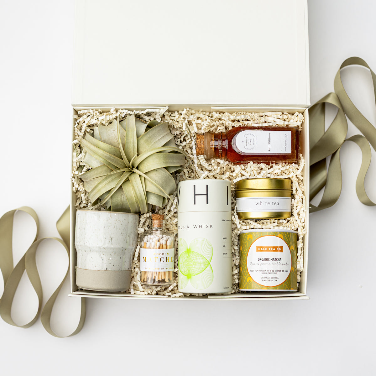A curated gift box set containing a ceramic candle, a small bottle of chamomile tea, a tin of white tea, a tin of blue raspberry lemonade, and a decorative dried palm leaf, arranged in shredded paper inside a white box with a ribbon.
