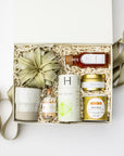 A curated gift box set containing a ceramic candle, a small bottle of chamomile tea, a tin of white tea, a tin of blue raspberry lemonade, and a decorative dried palm leaf, arranged in shredded paper inside a white box with a ribbon.