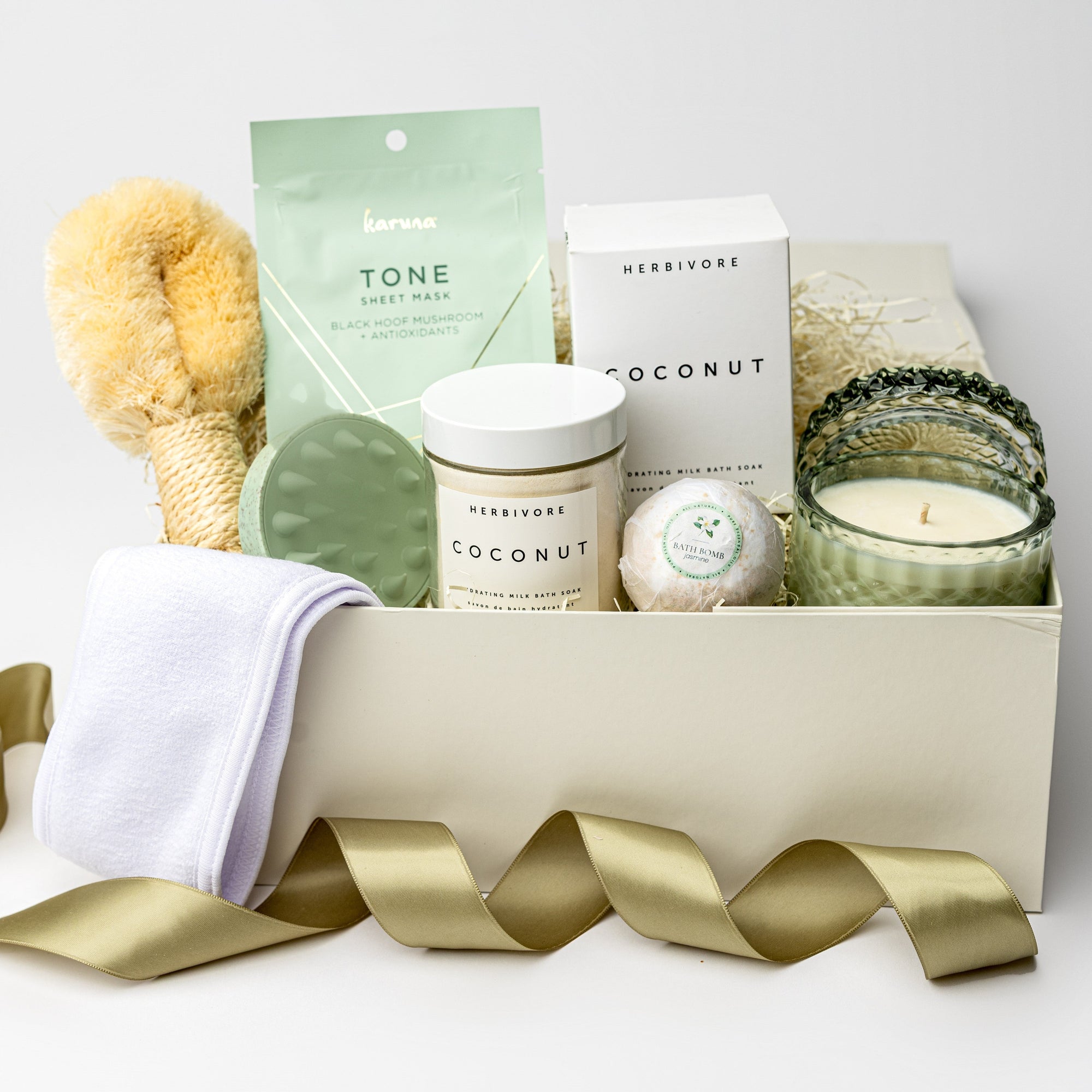 A front view of green goddess self-care gift set featuring a spa headband, a Tone face mask, a green scalp massager, a coconut milk soak, a jasmine bath bomb, a white tea trinket green jar candle, and a Nordic brush, all arranged in a white gift box with ivory shred and tied with a green ribbon on a white background.