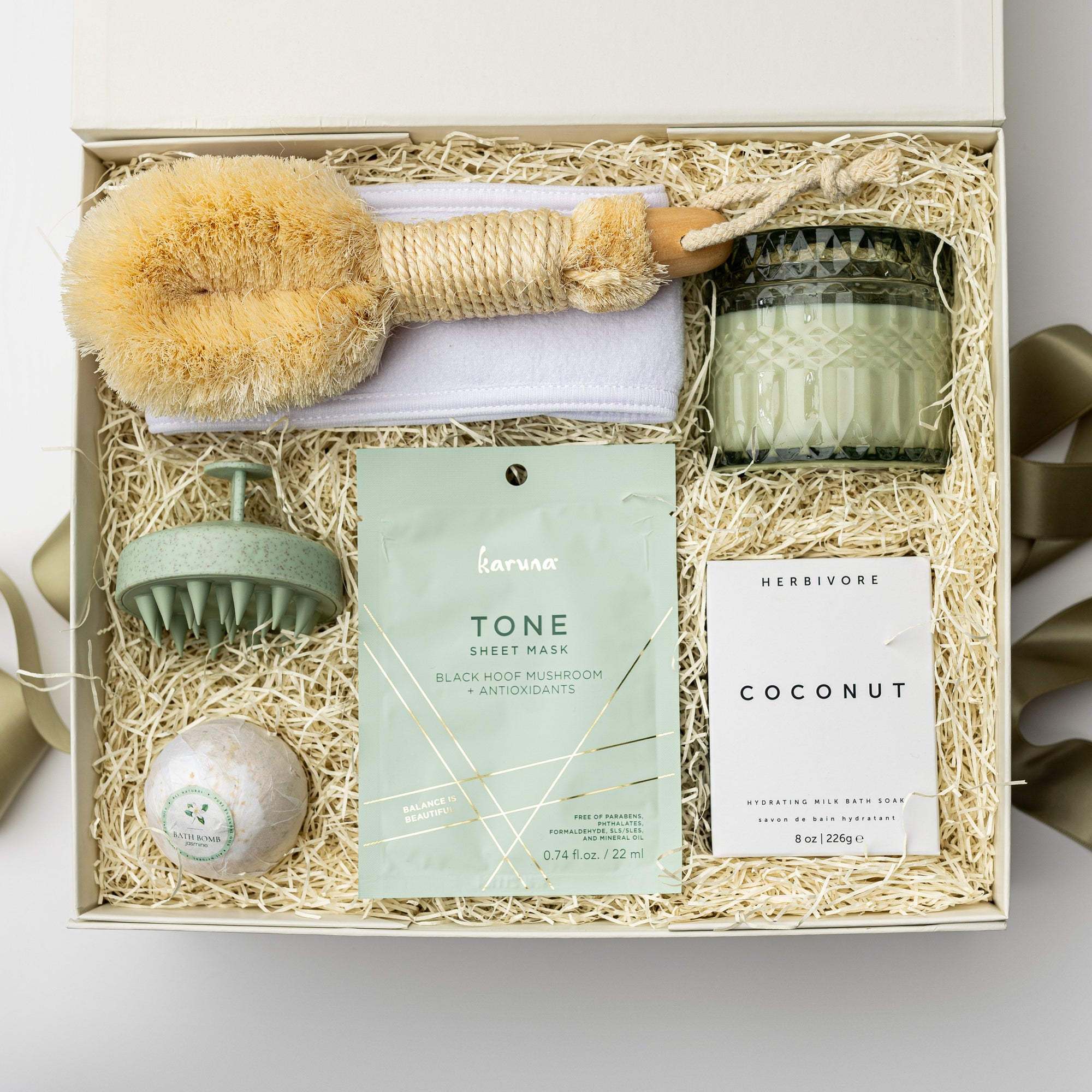 A top view of green goddess self-care gift set featuring a spa headband, a Tone face mask, a green scalp massager, a coconut milk soak, a jasmine bath bomb, a white tea trinket green jar candle, and a Nordic brush, all arranged in a white gift box with ivory shred and tied with a green ribbon on a white background.