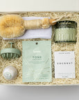 A top view of green goddess self-care gift set featuring a spa headband, a Tone face mask, a green scalp massager, a coconut milk soak, a jasmine bath bomb, a white tea trinket green jar candle, and a Nordic brush, all arranged in a white gift box with ivory shred and tied with a green ribbon on a white background.