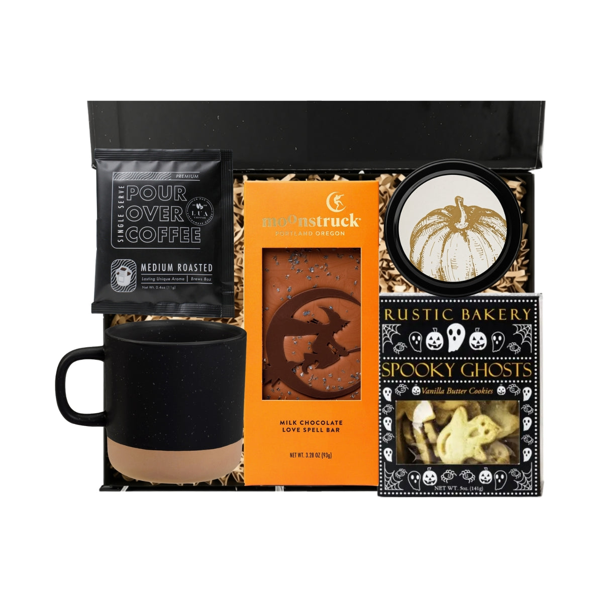 A Halloween-themed gift basket set containing a black mug with a beige base, a black pour-over coffee bag, an orange chocolate love feel bar, a pumpkin candle, and a Rustic Bakery Spooky Ghosts cookie tin, all arranged in a black box with shredded paper.