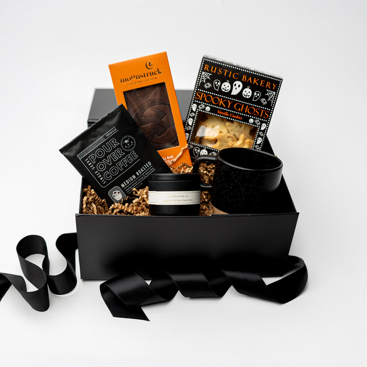 A curated gift box arrangement on a white background, featuring a black gift box filled with packaged snacks and a black speckled mug. Visible items include a bag of your toffee coffee, a jar of brown butter, a box of rustic baked sticky cookies, and an orange box of chocolate-covered treats, with black ribbon draped around the box.