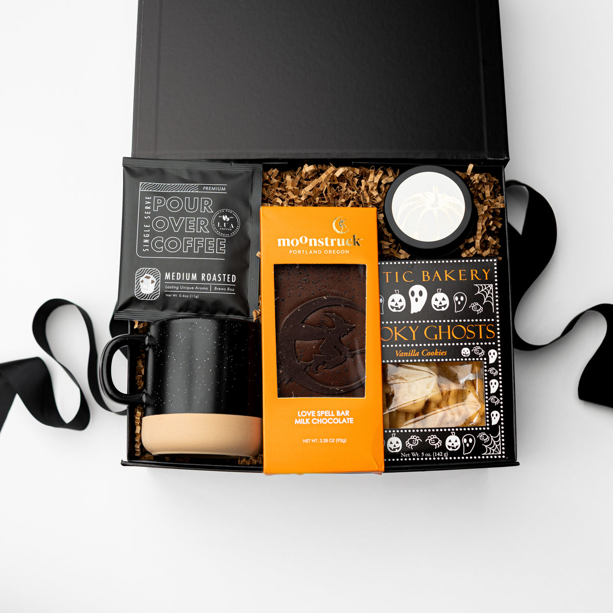 An open black gift box arranged on a white background with a black coffee mug, a bag of pour-over coffee, an orange package of mo'nsstruk chocolate, and a package of spooky themed cookies.